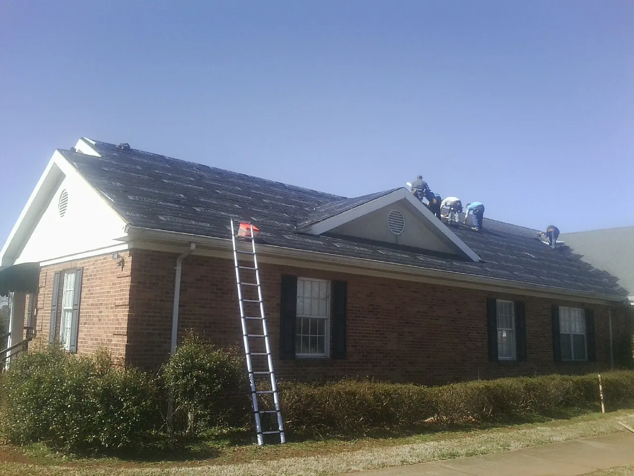 Crew tearing shingles off a brick ranch roof with a ladder set — Emergency Roof Repair work by our crew in Jeremiah