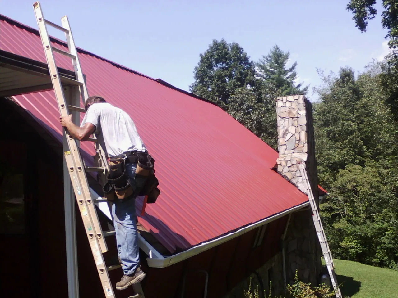 Roofer on a ladder beside a red metal roof and stone chimney — Soffit & Fascia Repair by Roofing Brothers in Jeremiah